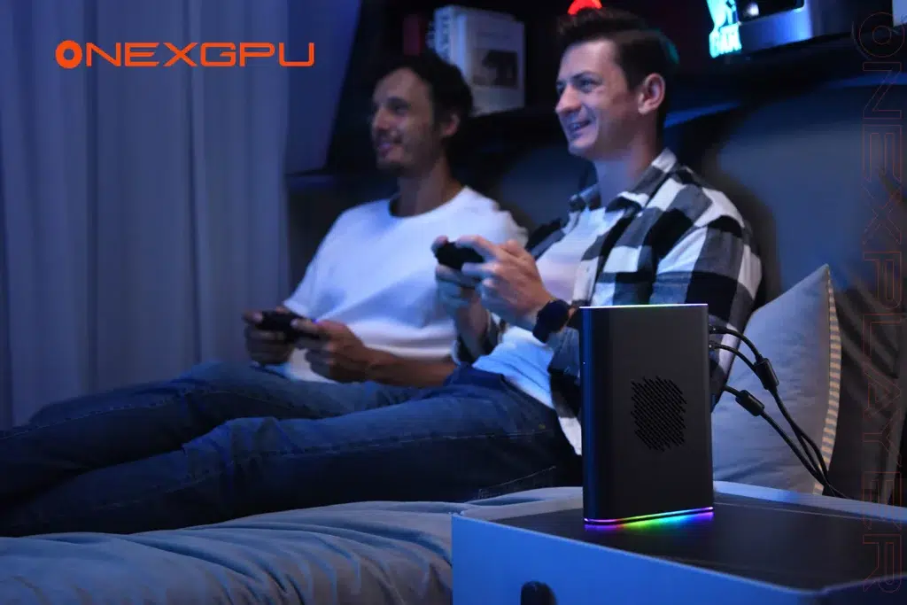 ONEXPLAYER ONEXGPU 2 eGPU Docking Station 1 Two men sitting on a couch gaming with controllers in hand, illuminated by ambient blue lighting. A sleek external GPU device, the ONEXGPU 2, sits on a nearby table, featuring RGB lighting and multiple connected cables. The ONEXGPU logo is prominently displayed.
