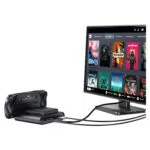 A black GPD WIN 5 handheld console is shown docked and connected via cables to a monitor The monitor displays the Steam OS style interface with a grid of game titles such as Dead Cells Deathloop Portal 2 and Stardew Valley illustrating the devices ability to drive a desktop gaming experience