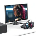 A desk setup showing the GPD WIN 5 docked and connected to an external monitor via cables The same image of a female character with pink hair is displayed on both the small console screen and the large external monitor demonstrating the docks video output capability